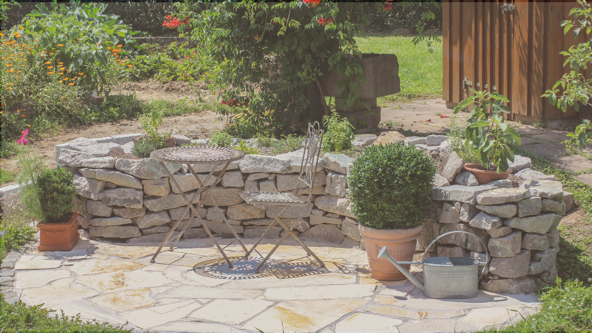 natürliches Hochbeet aus grauen Mauersteinen, neben einem kleinen Sitzbereich aus sandfarbenen Polygonalplatten