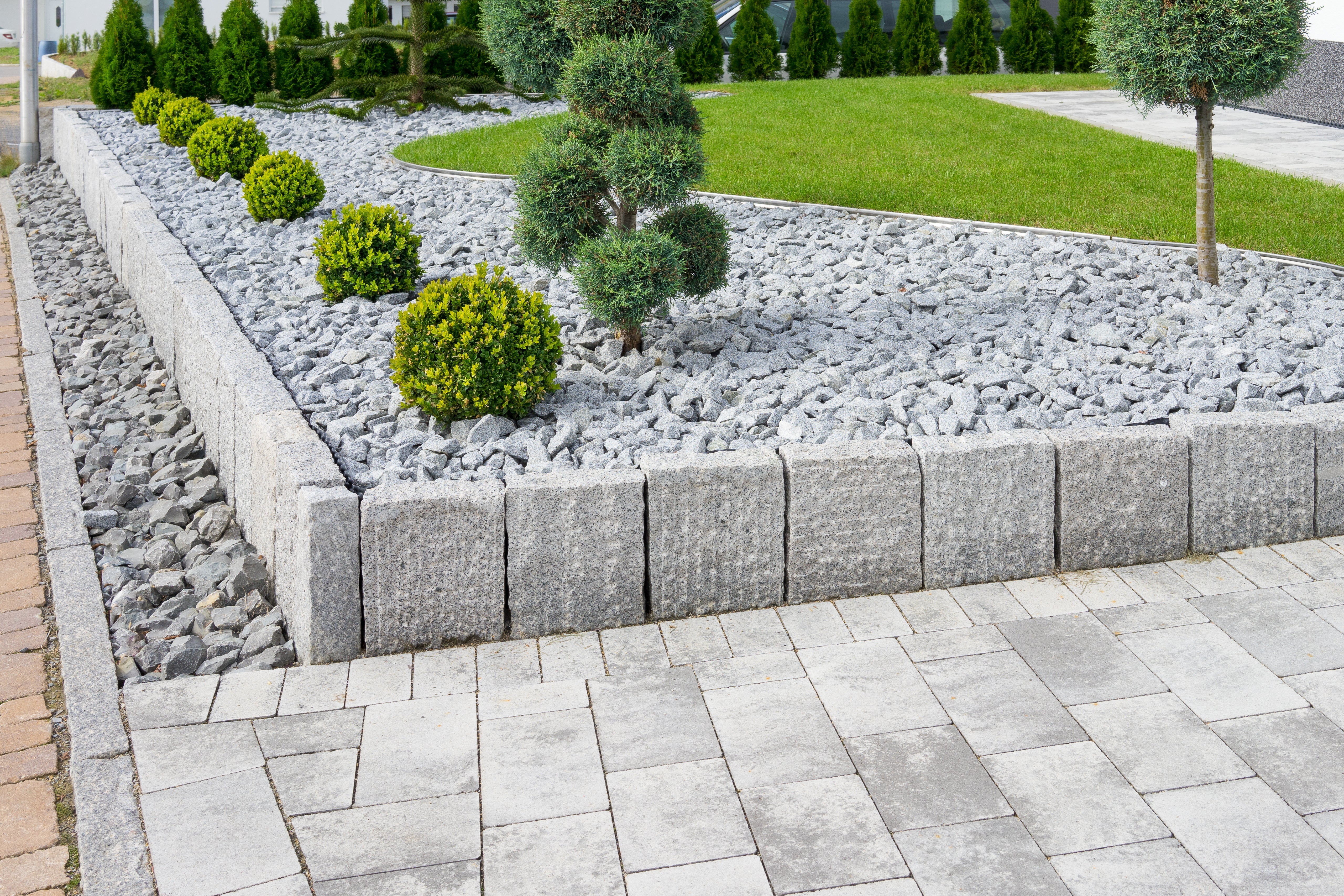 Garten mit Hochbeet aus Granit Palisaden. Das Beet ist mit Kies und kleinen Bäumen bepflanzt. Im Vordergrund ein Weg aus Natursteinplatten.
